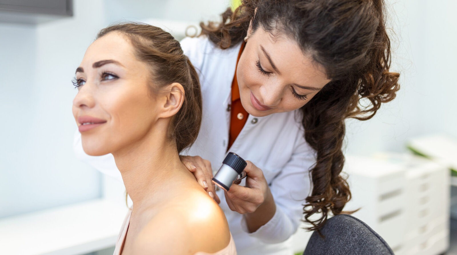 Female dermatologist carefully examining the skin of a female patient using a dermascope, looking for signs of skin cancer. Dermatologist examining patient's birthmark with magnifying glass in clinic